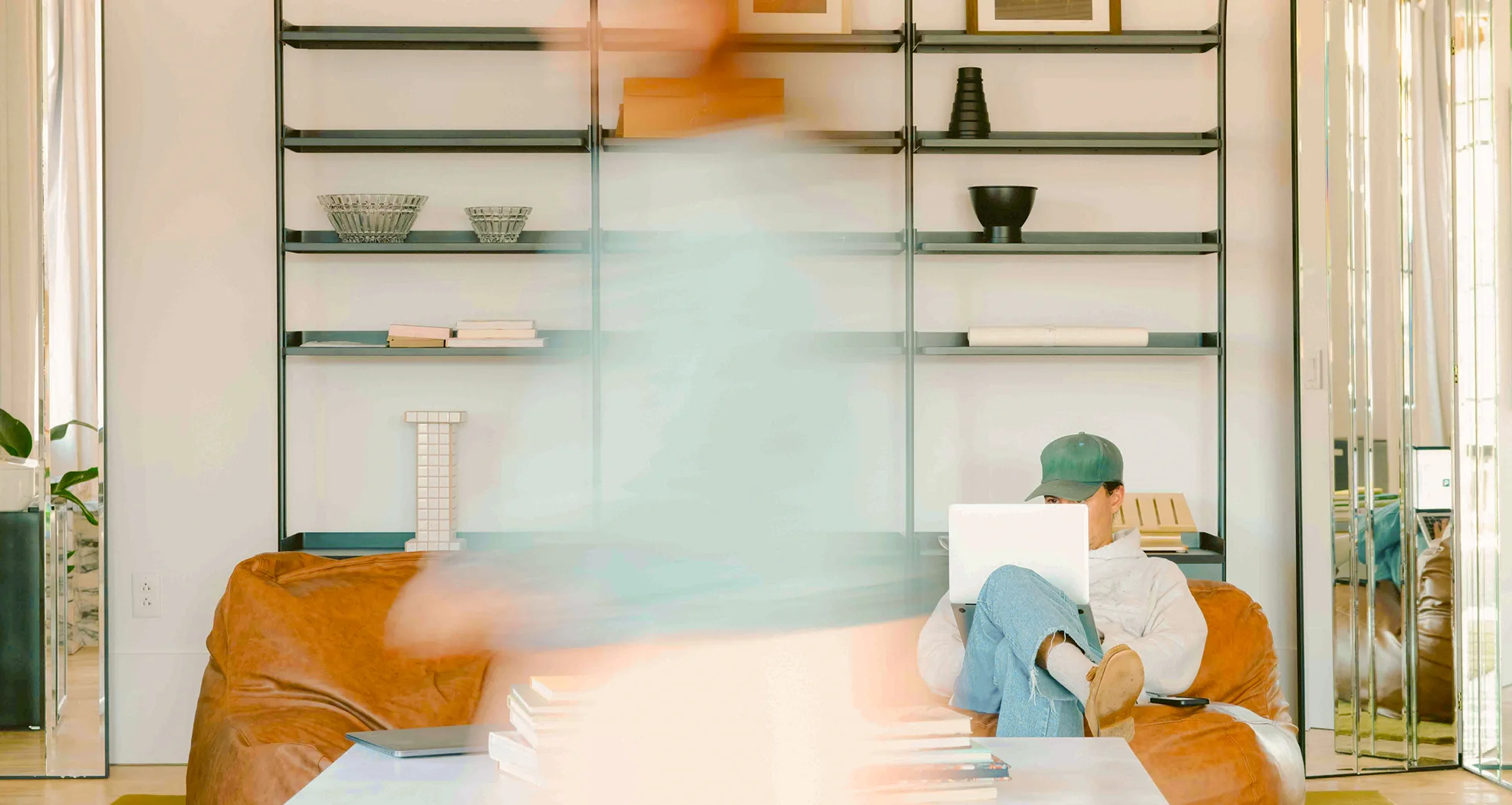 A blurred photo of a person walking through a room with someone working on a laptop.